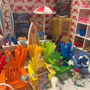 Puede incluir: Una escena de playa en miniatura con sillas Adirondack coloridas, tablas de surf y sombrillas. La escena incluye patos en miniatura, cubos y un letrero que dice "Maine Blueberries". El esquema de color es brillante y alegre.