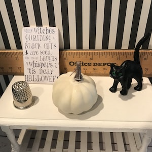May include: A Halloween-themed display featuring a white pumpkin, a black cat figurine with green eyes, a silver thimble, and a sign with the text "When witches go riding... 'tis near Halloween". The items are arranged on a white table.