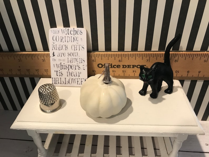 May include: A Halloween-themed display featuring a white pumpkin, a black cat figurine with green eyes, a silver thimble, and a sign with the text "When witches go riding... 'tis near Halloween". The items are arranged on a white table.