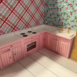 May include: A pink miniature kitchen playset with a white countertop, a stovetop with four burners, an oven, a sink, and cabinets. The kitchen is made of wood and has a white countertop.