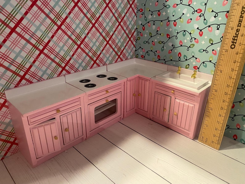 May include: A pink miniature kitchen playset with a white countertop, a stovetop with four burners, an oven, a sink, and cabinets. The kitchen is made of wood and has a white countertop.