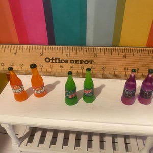 May include: A collection of miniature soda bottles in orange, green, and purple. The orange bottles are labelled "Crush", the green bottles have "Mountain Dew" labels, and the purple bottles have labels with a black and white design. The bottles are on a white table.