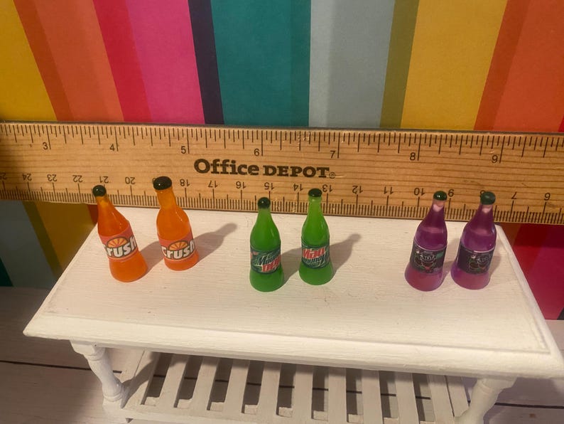 May include: A collection of miniature soda bottles in orange, green, and purple. The orange bottles are labelled "Crush", the green bottles have "Mountain Dew" labels, and the purple bottles have labels with a black and white design. The bottles are on a white table.