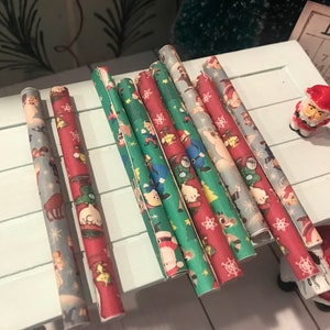 Puede incluir: Surtido de ocho rollos de papel de regalo de Navidad con varios dise&ntilde;os de tem&aacute;tica navide&ntilde;a que incluyen Pap&aacute; Noel, renos y copos de nieve. El papel es rojo, verde y azul con detalles blancos.