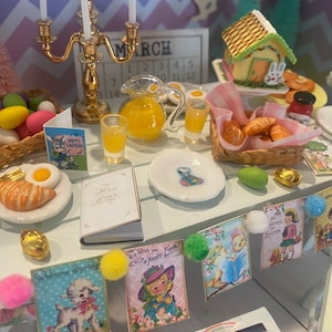 May include: A miniature Easter table setting with a white tablecloth, a pitcher of orange juice, a plate of croissants, a plate of eggs, and a plate with a fried egg and a croissant. There are also several Easter-themed cards and decorations on the table.