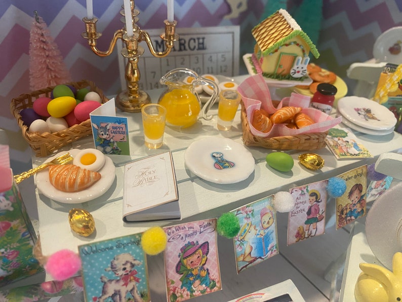 May include: A miniature Easter table setting with a white tablecloth, a pitcher of orange juice, a plate of croissants, a plate of eggs, and a plate with a fried egg and a croissant. There are also several Easter-themed cards and decorations on the table.