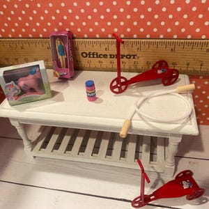 Op de afbeelding: Miniatuurspeelgoed, waaronder een roze pony, een pop, een rode step, een springtouw en een flesje bellenblaas, staan op een witte tafel. Op de achtergrond een liniaal en een rood-wit stippenpatroon.