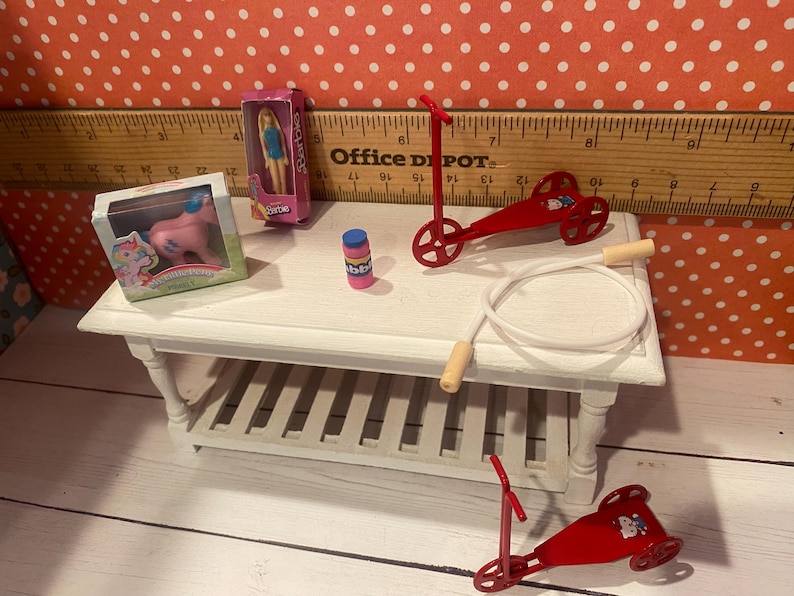 Op de afbeelding: Miniatuurspeelgoed, waaronder een roze pony, een pop, een rode step, een springtouw en een flesje bellenblaas, staan op een witte tafel. Op de achtergrond een liniaal en een rood-wit stippenpatroon.