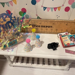 May include: A miniature white table displays birthday party decorations. There are three party hats with colorful polka dots and pom-poms, a small black camera, and a box game. A glittery "Happy Birthday" sign is also present.