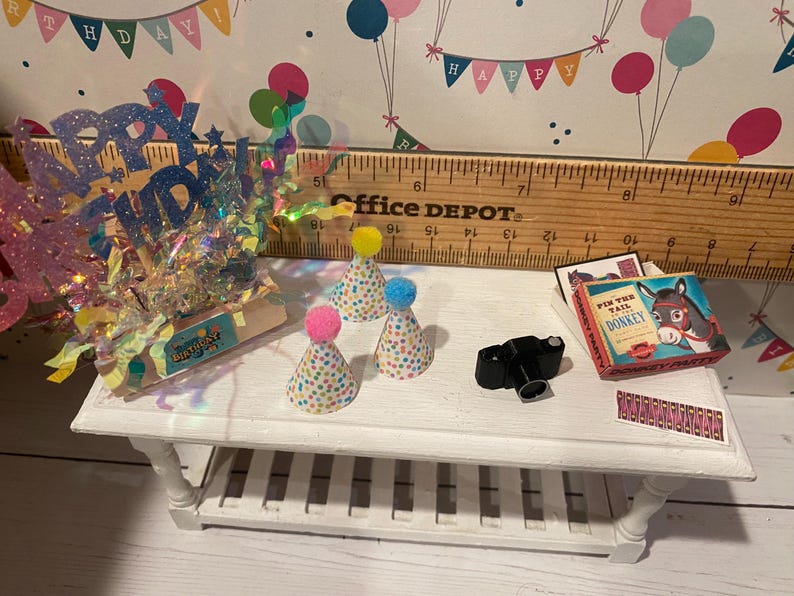 May include: A miniature white table displays birthday party decorations. There are three party hats with colorful polka dots and pom-poms, a small black camera, and a box game. A glittery "Happy Birthday" sign is also present.