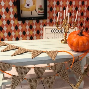 May include: A miniature table setting with a burlap banner, a gold candlestick with white candles, a small orange pumpkin, and a framed picture of a ghost reading a book. The table has a light blue surface and the background is a diamond pattern wallpaper with orange, yellow, and black colors. The calendar on the table says "October" with the numbers 6, 7, 8, 9, 1, 2.