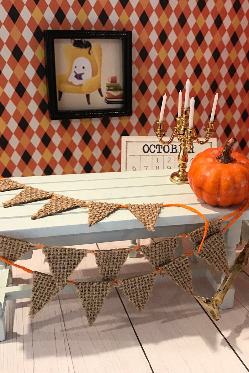 May include: A miniature table setting with a burlap banner, a gold candlestick with white candles, a small orange pumpkin, and a framed picture of a ghost reading a book. The table has a light blue surface and the background is a diamond pattern wallpaper with orange, yellow, and black colors. The calendar on the table says "October" with the numbers 6, 7, 8, 9, 1, 2.