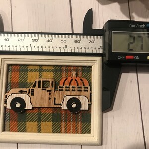 May include: A wooden cutout of a truck carrying a pumpkin, framed in a white frame with a plaid fabric background. The frame measures 2.71 inches in width.