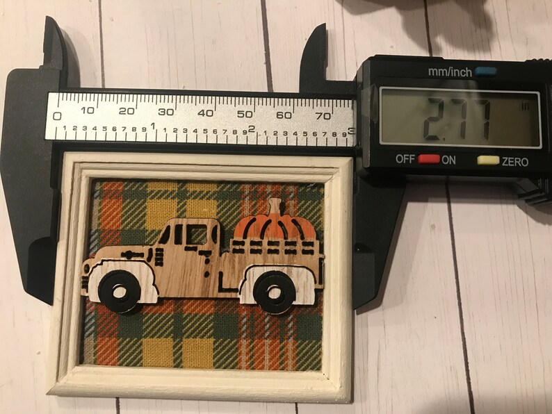 May include: A wooden cutout of a truck carrying a pumpkin, framed in a white frame with a plaid fabric background. The frame measures 2.71 inches in width.