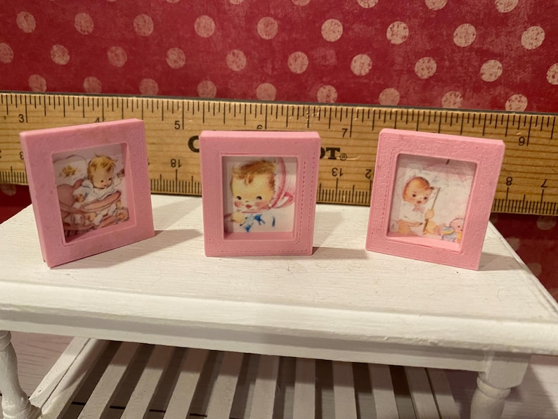 Puede incluir: Tres peque&ntilde;as im&aacute;genes enmarcadas en rosa con ilustraciones de beb&eacute;s vintage. Los marcos son cuadrados y est&aacute;n sobre una mesa blanca. Una regla de madera est&aacute; en el fondo. Las im&aacute;genes de beb&eacute;s son en colores pastel suaves.