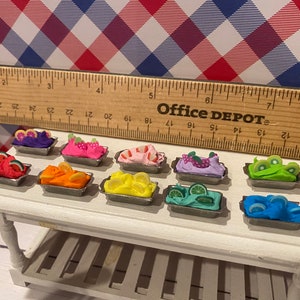 May include: Miniature fruit-topped desserts in small metal trays. The desserts feature watermelon, orange slices, lemon, kiwi, and other fruits. The desserts are arranged on a white table with a ruler and a red, white, and blue checkered background.