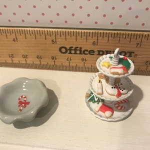 May include: A white ceramic three-tiered serving stand with a variety of Christmas-themed cookies, including a snowman, a stocking, and a candy cane. A small white ceramic bowl with a red and white candy cane design is also pictured.