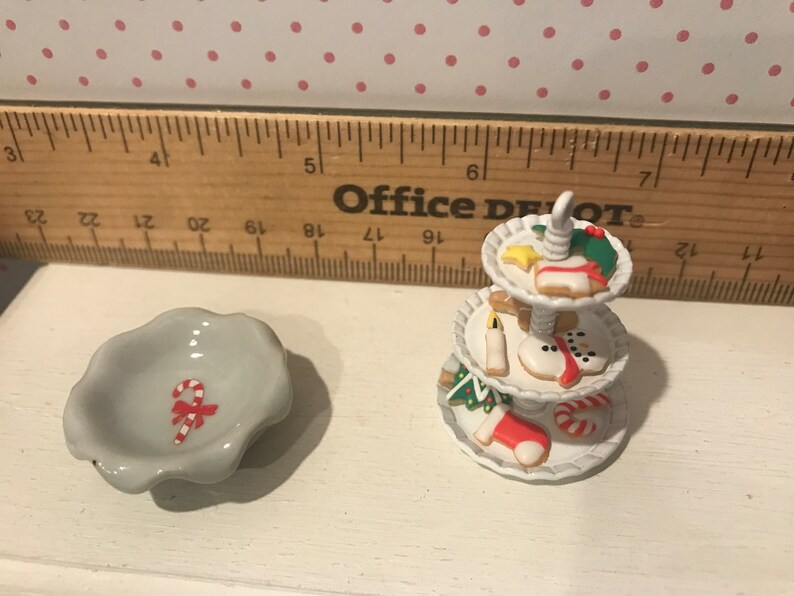 May include: A white ceramic three-tiered serving stand with a variety of Christmas-themed cookies, including a snowman, a stocking, and a candy cane. A small white ceramic bowl with a red and white candy cane design is also pictured.