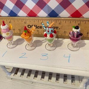 May include: Four miniature sundae desserts in clear glass cups. Each sundae has different toppings and colors, including pink, orange, blue, and purple. The desserts are arranged on a small white table with a ruler in the background.