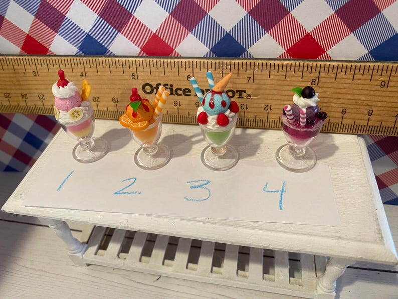 May include: Four miniature sundae desserts in clear glass cups. Each sundae has different toppings and colors, including pink, orange, blue, and purple. The desserts are arranged on a small white table with a ruler in the background.