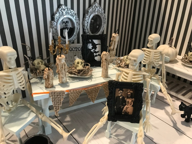 May include: A Halloween-themed miniature dining scene with skeleton figures seated around a white table. The table is decorated with candles, skulls, and a burlap banner. A framed vintage photograph and a black cat figurine add to the spooky ambiance. The background features black and white striped wallpaper.