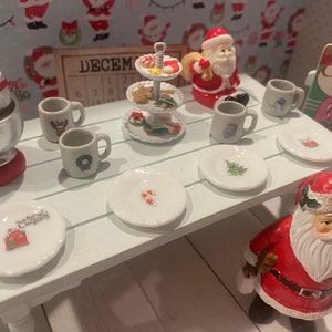 May include: A white table with a white and red candy cane design on a plate. There are six white plates with different Christmas designs on them. There are four white mugs with Christmas designs on them. There is a three-tiered white serving tray with Christmas cookies and candy on it. There is a silver stand mixer with a red base. There is a Santa Claus figurine holding a bag of gifts. There is a sign that says "It's a Merry Snowy Candy Canes".