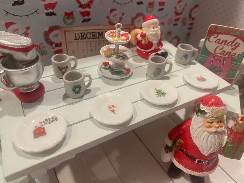 May include: A white table with a white and red candy cane design on a plate. There are six white plates with different Christmas designs on them. There are four white mugs with Christmas designs on them. There is a three-tiered white serving tray with Christmas cookies and candy on it. There is a silver stand mixer with a red base. There is a Santa Claus figurine holding a bag of gifts. There is a sign that says "It's a Merry Snowy Candy Canes".