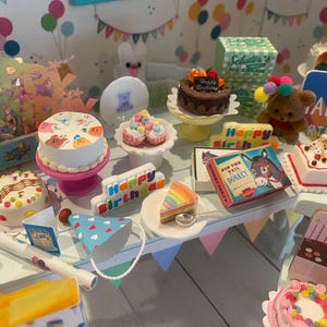 May include: A collection of miniature birthday-themed items, including cakes, party hats, and a teddy bear. The scene includes a cake with the words "Happy Birthday" and a vintage "Pin the Tail on the Donkey" game.