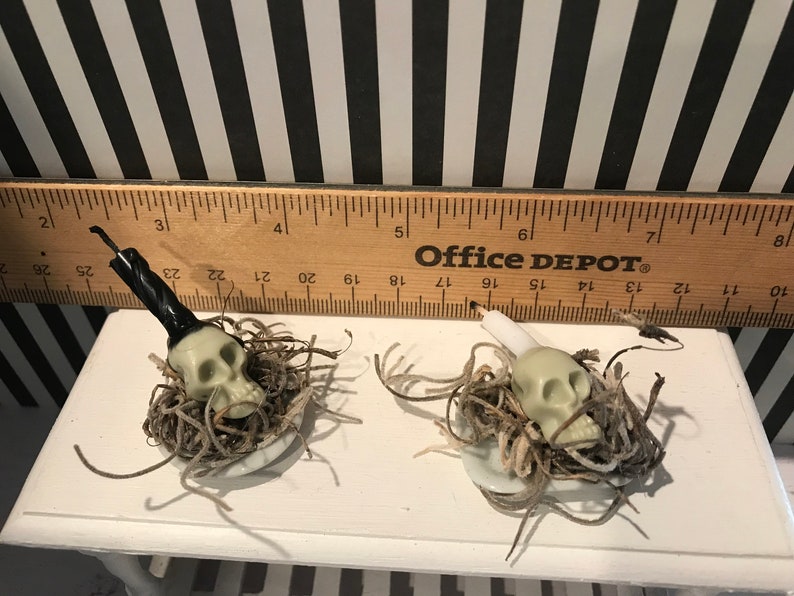 May include: Two small Halloween skull decorations. Each skull is nestled in a bed of brown and tan twine, sitting on a small white plate. One skull has a black candle, the other has a white candle. A wooden ruler is in the background.