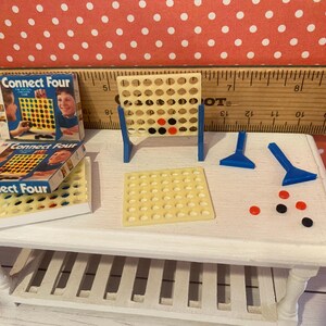 Op de afbeelding: Miniatuur Connect Four-spelset met een beige spelbord, blauwe steunen, spelstukken en twee dozen. De dozen zijn blauw en wit met de spelnaam. Rode, zwarte en beige spelstukken liggen op de tafel.
