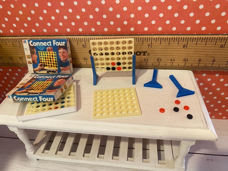 Op de afbeelding: Miniatuur Connect Four-spelset met een beige spelbord, blauwe steunen, spelstukken en twee dozen. De dozen zijn blauw en wit met de spelnaam. Rode, zwarte en beige spelstukken liggen op de tafel.