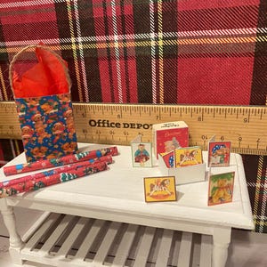 Puede incluir: Una escena navide&ntilde;a en miniatura con una bolsa de regalo azul con hombres de jengibre, rollos de papel de regalo rojos y peque&ntilde;as tarjetas. Las tarjetas muestran varias ilustraciones navide&ntilde;as. La escena est&aacute; ambientada sobre una mesa blanca.