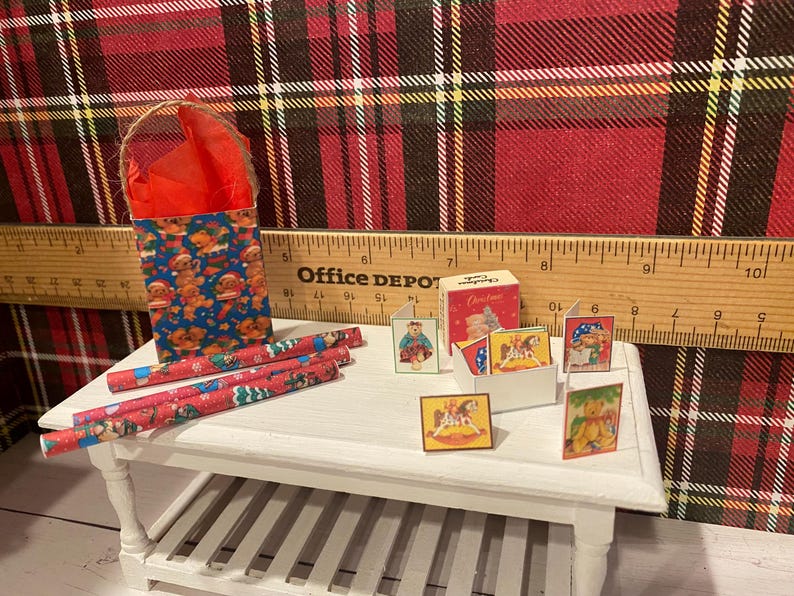 Puede incluir: Una escena navide&ntilde;a en miniatura con una bolsa de regalo azul con hombres de jengibre, rollos de papel de regalo rojos y peque&ntilde;as tarjetas. Las tarjetas muestran varias ilustraciones navide&ntilde;as. La escena est&aacute; ambientada sobre una mesa blanca.