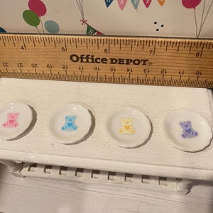 May include: Four small, white, scalloped-edge plates with colorful teddy bear designs. The bears are pink, blue, yellow, and purple. The plates are arranged on a white, decorative table. A wooden ruler is above the plates.
