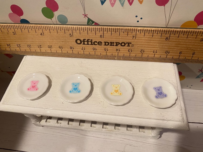 May include: Four small, white, scalloped-edge plates with colorful teddy bear designs. The bears are pink, blue, yellow, and purple. The plates are arranged on a white, decorative table. A wooden ruler is above the plates.