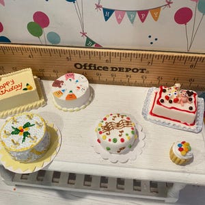 May include: A collection of miniature cakes, including a rectangular cake with "Happy Birthday" text, a round cake with animal faces, and a cupcake. The cakes are decorated with various toppings and sit on a white surface.