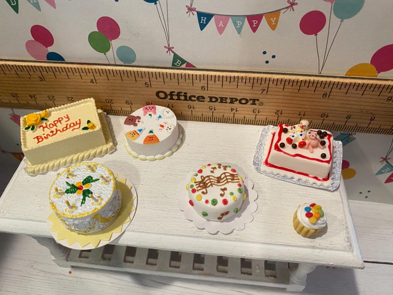 May include: A collection of miniature cakes, including a rectangular cake with "Happy Birthday" text, a round cake with animal faces, and a cupcake. The cakes are decorated with various toppings and sit on a white surface.