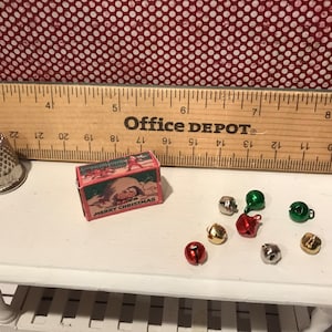 May include: A miniature Christmas scene featuring a thimble, a small box with "MERRY CHRISTMAS" text, and several tiny bells in various colors. A wooden ruler is in the background.