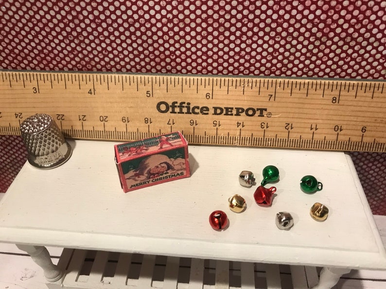 May include: A miniature Christmas scene featuring a thimble, a small box with "MERRY CHRISTMAS" text, and several tiny bells in various colors. A wooden ruler is in the background.
