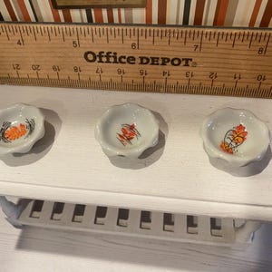 May include: Three small, white, scalloped-edge ceramic dishes with a floral design in orange, black, and yellow. The dishes are arranged on a white shelf, with a wooden ruler and the text "Office DEPOT" in the background.