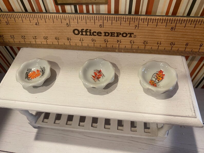 May include: Three small, white, scalloped-edge ceramic dishes with a floral design in orange, black, and yellow. The dishes are arranged on a white shelf, with a wooden ruler and the text "Office DEPOT" in the background.
