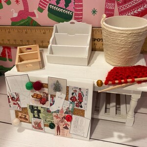 May include: Miniature white desk with a corkboard, small wooden drawer, and a three-tiered white organizer. A woven basket, red yarn, and knitting needles are also on the desk. The corkboard displays various vintage-style images and is decorated with colorful pom-poms.