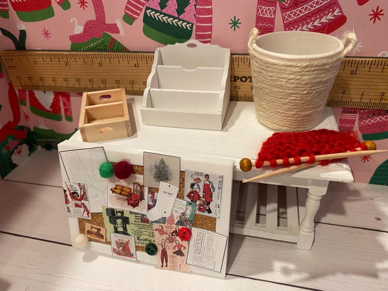 May include: Miniature white desk with a corkboard, small wooden drawer, and a three-tiered white organizer. A woven basket, red yarn, and knitting needles are also on the desk. The corkboard displays various vintage-style images and is decorated with colorful pom-poms.