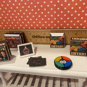 Op de afbeelding: Miniatuur vintage speelgoed, waaronder Lite-Brite en Simon speldozen, een Lite-Brite scherm met een clown ontwerp en een Simon spelunit. Het speelgoed is gerangschikt op een witte tafel.