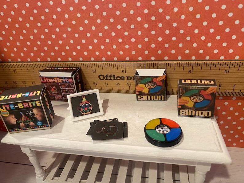 Op de afbeelding: Miniatuur vintage speelgoed, waaronder Lite-Brite en Simon speldozen, een Lite-Brite scherm met een clown ontwerp en een Simon spelunit. Het speelgoed is gerangschikt op een witte tafel.