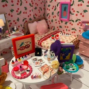Puede incluir: Una escena de dormitorio en miniatura con papel pintado con estampado de cerezas, una cama con almohadas rosas y un tocador con espejo. Una pequeña mesa exhibe maquillaje, esmalte de uñas y obras de arte enmarcadas. La habitación está decorada en tonos rosa, blanco y rojo.