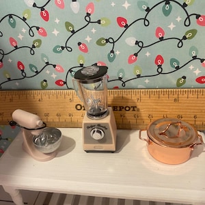 May include: A set of three miniature kitchen appliances in a cream and black color scheme. The appliances include a blender, a stand mixer, and a copper pot with a lid.