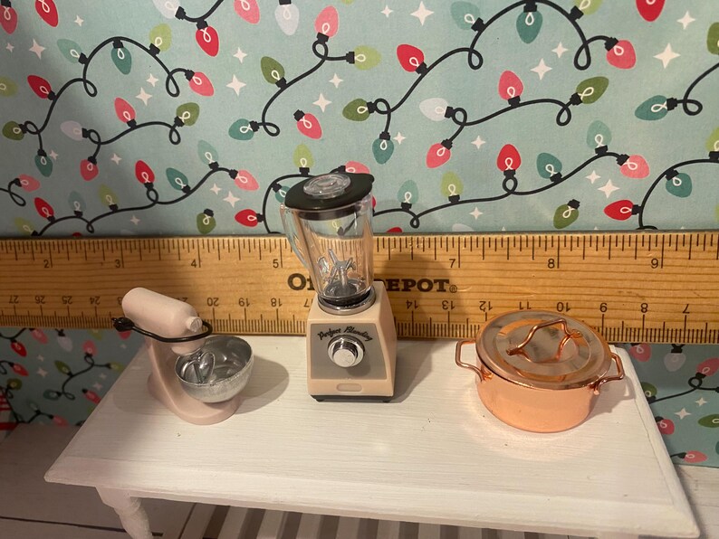 May include: A set of three miniature kitchen appliances in a cream and black color scheme. The appliances include a blender, a stand mixer, and a copper pot with a lid.
