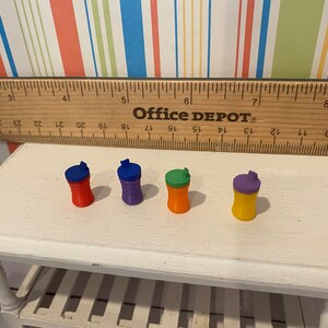 May include: Four miniature, colorful water bottles with flip-top lids are arranged on a white table. The bottles are red, purple, orange, and yellow, each with a contrasting lid color. A wooden ruler and the text "Office DEPOT" are in the background.