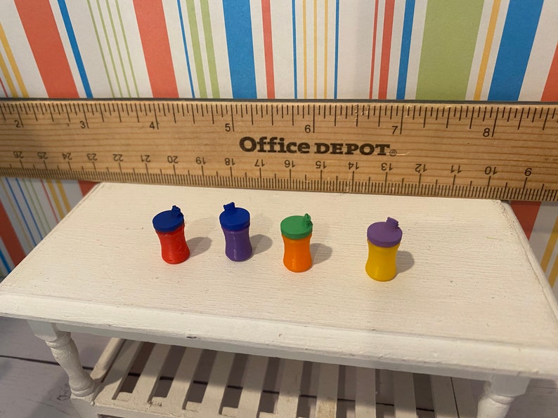 May include: Four miniature, colorful water bottles with flip-top lids are arranged on a white table. The bottles are red, purple, orange, and yellow, each with a contrasting lid color. A wooden ruler and the text "Office DEPOT" are in the background.